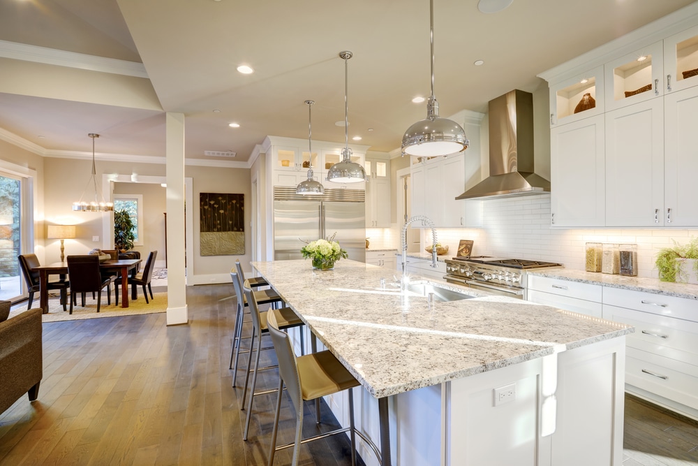 White kitchen design features large bar style kitchen island with granite countertop illuminated by modern pendant lights. Northwest, USA